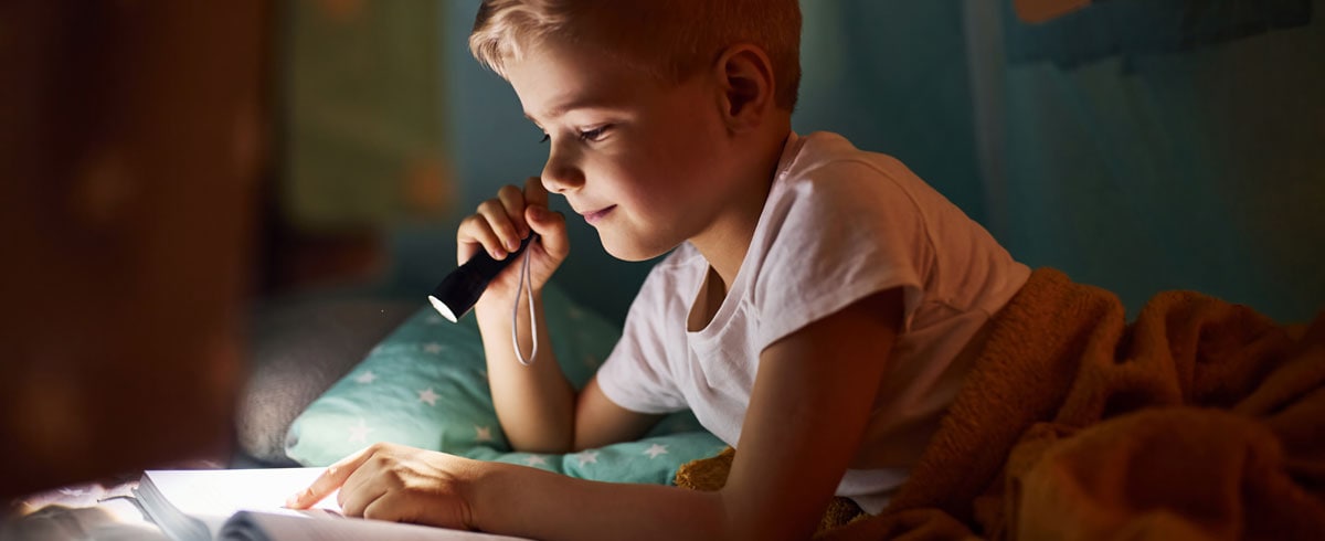Un bambino che legge un libro con una torcia elettrica in mano mentre è nel letto con una coperta