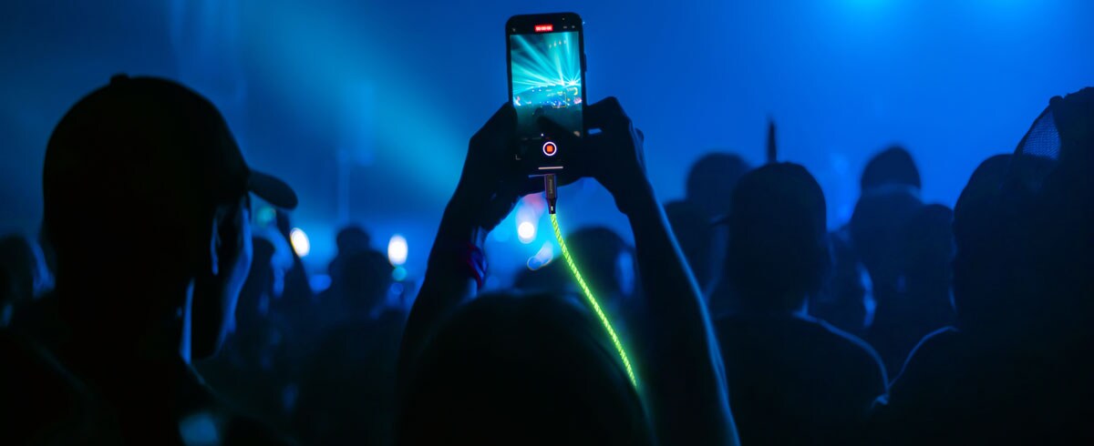 Una persona solleva un telefono cellulare collegato a un cavo luminoso di fronte a una folla, per catturare il momento durante un evento