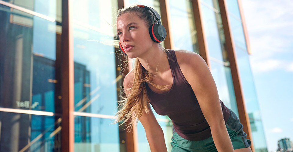 Una donna che indossa le cuffie Philips A6219 si prende una pausa durante un allenamento all'aperto, in piedi di fronte a un moderno edificio in vetro