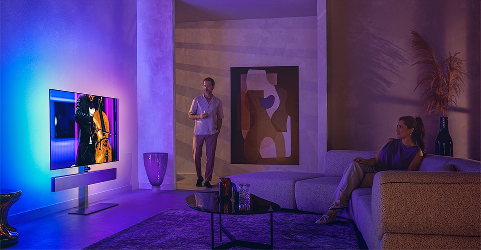 Un uomo e una donna in un elegante soggiorno guardano un televisore Philips Ambilight che trasmette un'esibizione di violoncello