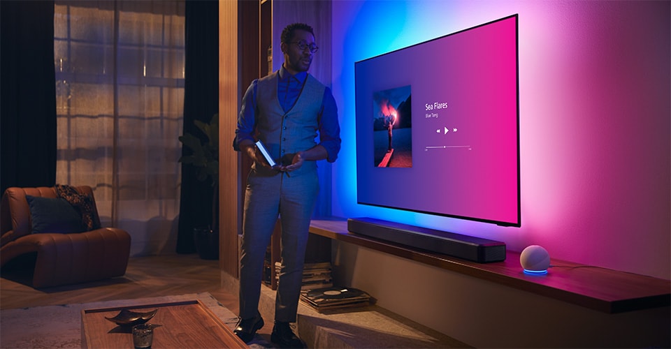 Persona in un soggiorno moderno vicino a una TV Philips Ambilight con interfaccia musicale e soundbar Fidelio