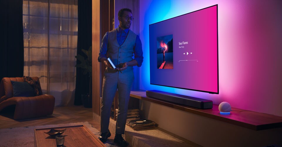 Persona in un soggiorno moderno vicino a una TV Philips Ambilight con interfaccia musicale e soundbar Fidelio