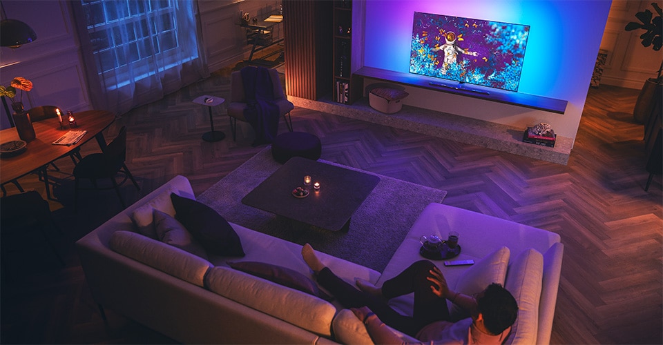 Uomo che si rilassa su un divano in un soggiorno scarsamente illuminato, guardando una TV Philips Ambilight