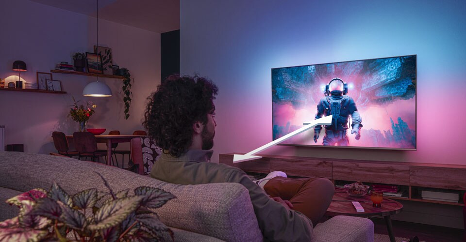 Uomo seduto su un divano che guarda una TV Philips Ambilight che mostra una scena colorata di un astronauta, con una freccia che indica la distanza di visione ottimale