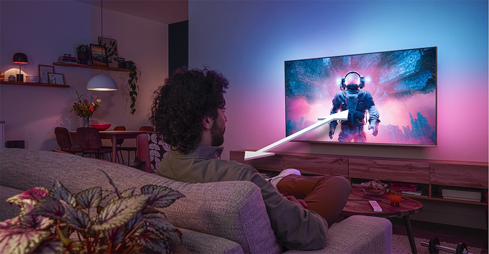 Uomo seduto su un divano che guarda una TV Philips Ambilight che mostra una scena colorata di un astronauta, con una freccia che indica la distanza di visione ottimale