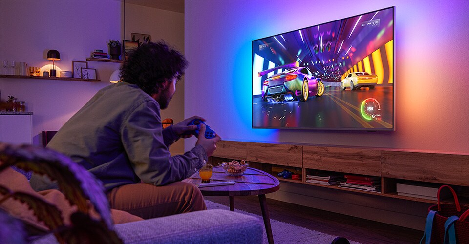  Una persona è seduta su un divano e gioca a un videogioco di corse su una TV Philips Ambilight