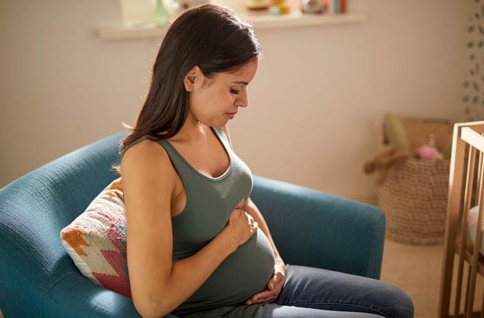 Una donna incinta è seduta sul divano con gli occhi chiusi, tenendosi teneramente la pancia.