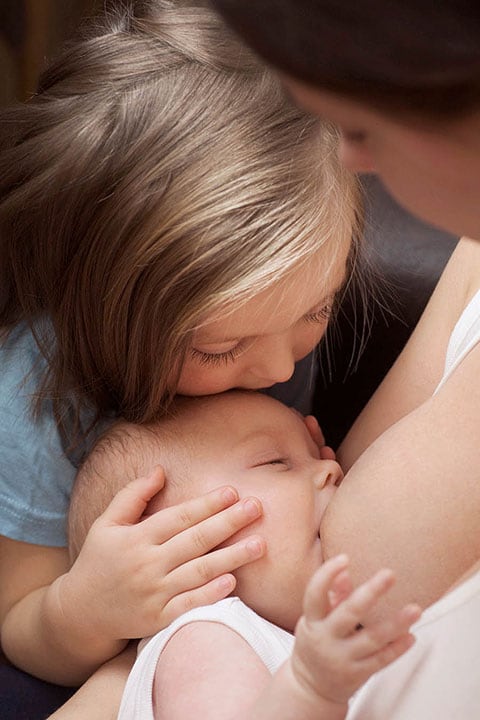 A mother breastfeeding her baby while her daughter holds the baby&#039;s head