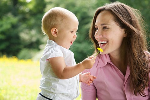 Mamma sorridente in un prato guarda con amore la sua bimba con un fiorellino in mano