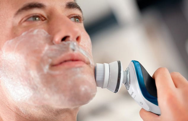 Primo piano di un uomo con la schiuma da barba sul viso che usa un rasoio elettrico bianco, blu e grigio.