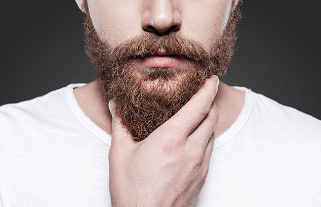 Primo piano del mento di un uomo con una maglietta bianca che tocca la sua folta barba marrone.