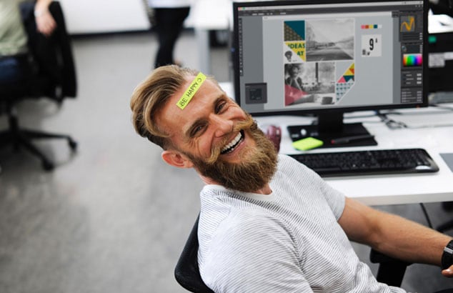 Ragazzo biondo, giovane e simpatico con una barba lunga è seduto su una sedia da ufficio e sorride davanti a sé.