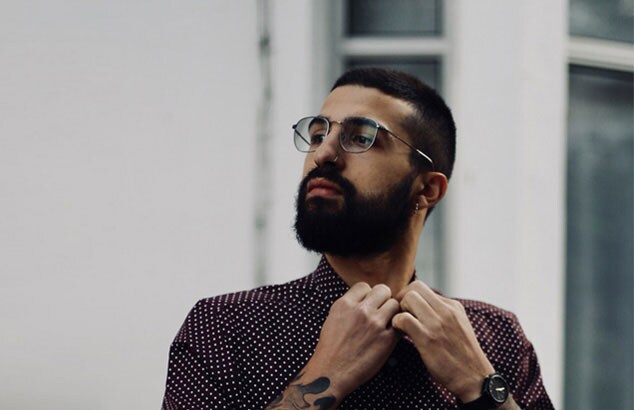 Un uomo con barba folta e scura, anello al naso e occhiali si abbottona la camicia rosso scuro a pois. 