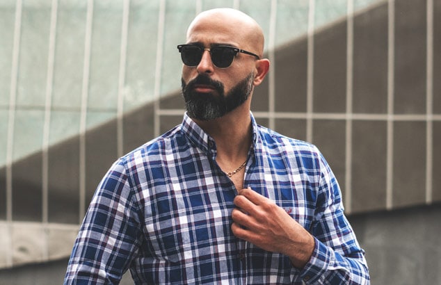 Elegante uomo calvo con una barba ben curata indossa una camicia a scacchi blu e occhiali da sole scuri.