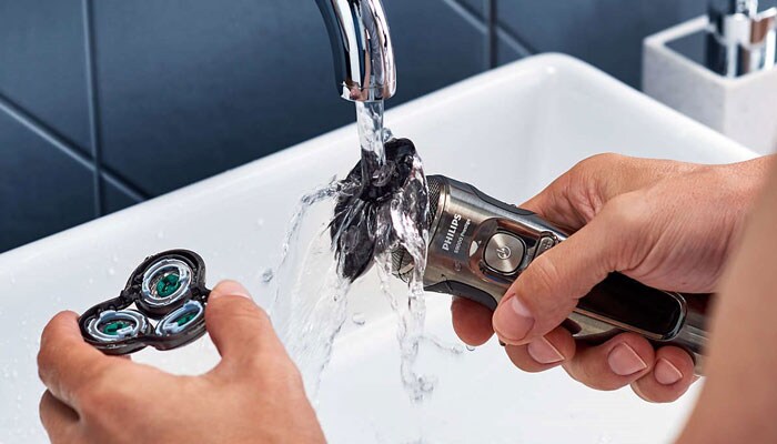 primo piano di una persona che tiene due parti di un rasoio elettrico nelle mani sotto il rubinetto dell'acqua.