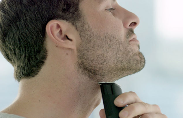 un uomo di profilo con la barba corta che si rade il collo con un rasoio elettrico.