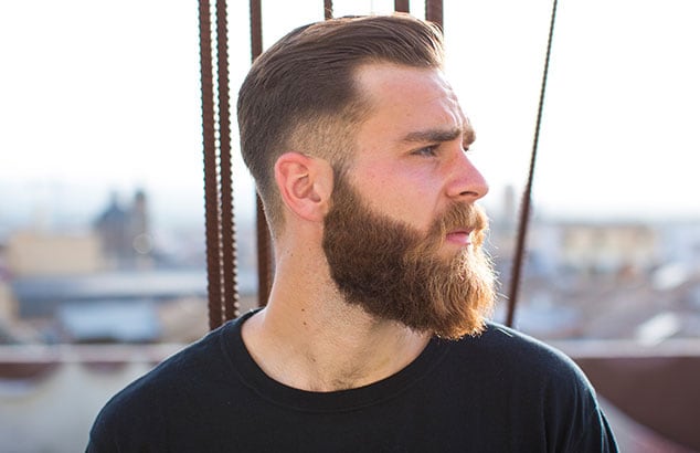 uomo di profilo con una lunga barba castana e capelli corti castani con una città sfocata sullo sfondo.