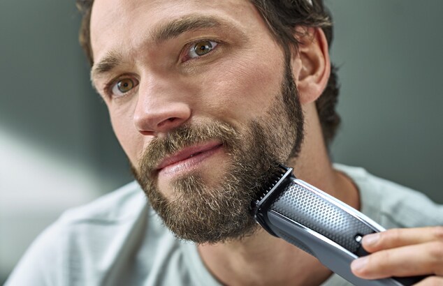 Un uomo con la barba folta si rade il viso con un rasoio elettrico argento e nero.