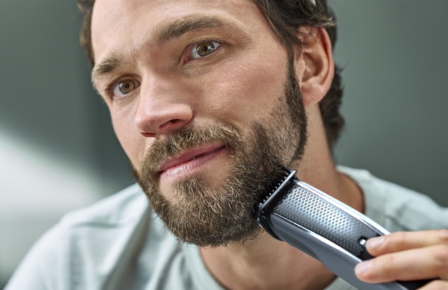 Primo piano di un uomo con la barba incolta che si rade il viso con un rasoio elettrico argento e nero.