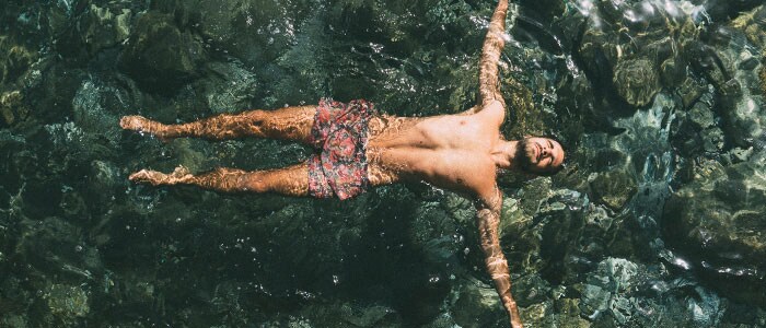 Foto dall’alto di un uomo castano con barba di media lunghezza in costume da bagno che galleggia in mare.
