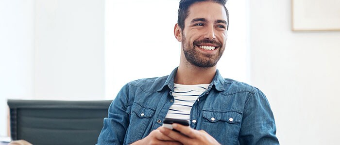 Primo piano di un uomo sorridente con barba incolta che indossa una maglietta a righe e ha in mano un telefono.